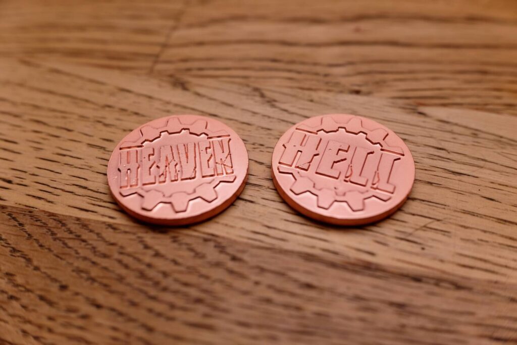 The health markers for each player are metal coins with the words "Heaven" and "Hell" engraved into them.