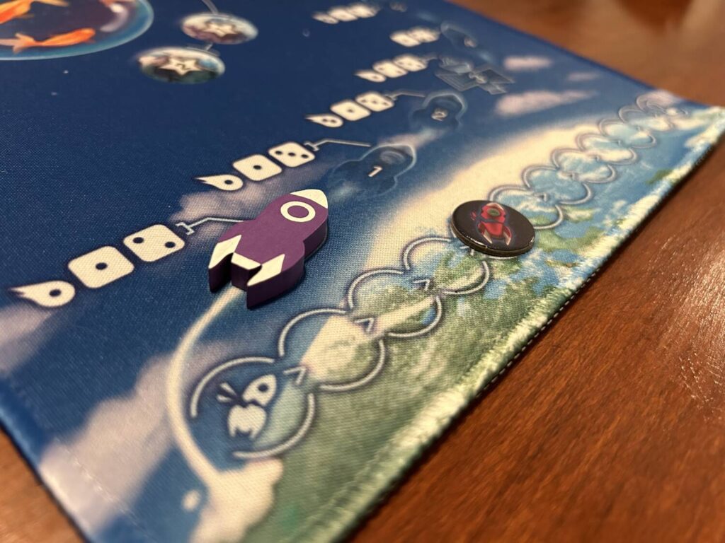 The rocket-shaped wooden token that tracks the mission's progress on the large neoprene mat, next to the cardboard token that tracks the number of failed missions.
