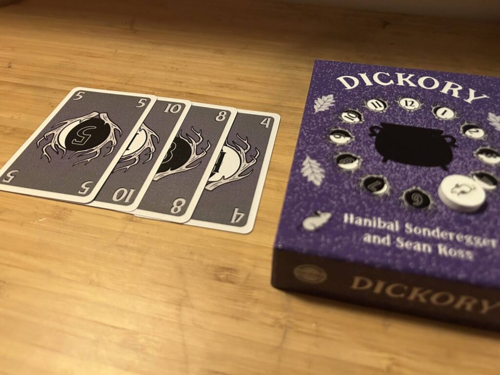 The Queue, a set of cards splayed out so all of them are visible but only one is fully so. I'll explain what the Queue is in a moment. Next to the Queue is the box for Dickory, which is used during gameplay. It's all very purple.