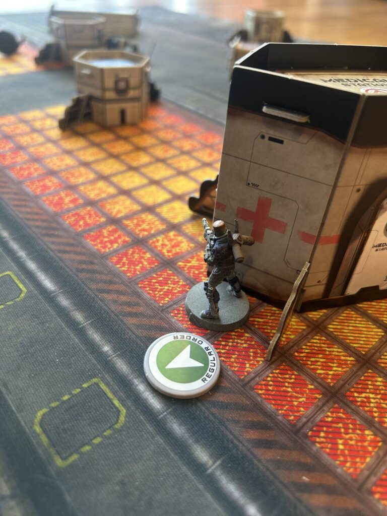 A figure stands next to flatpack terrain, an order token at its feet.