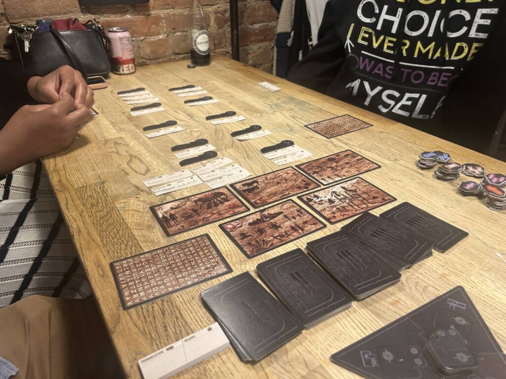 A wooden table, with two columns of small sheets of paper above multiple decks of cards.