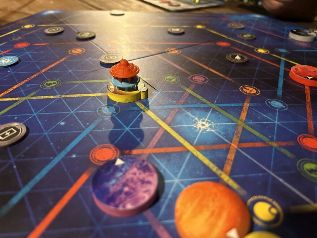 A close-up photograph of two plastic UFOs stacked on top of a wooden planet. The board underneath is dark blue, with paths in several colors running along it.