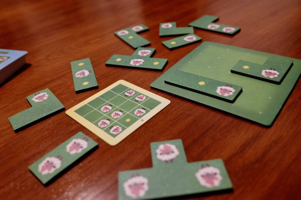 One of the puzzle cards next to the board and surrounded by the tetrominoes. Each piece has a green background, similar to a meadow, with sheep on top.