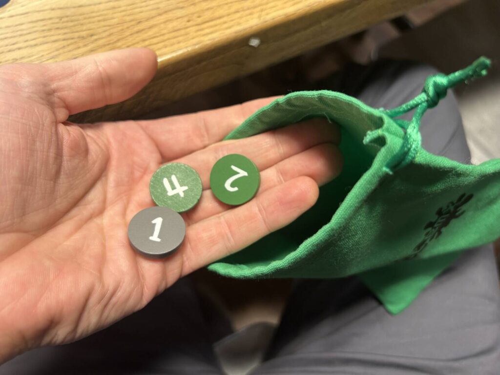 A hand emerges from a bag with three wooden tokens. Two of them, a 4 and a 2, are green. The third, a lowly 1 exhaustion token, is grey.