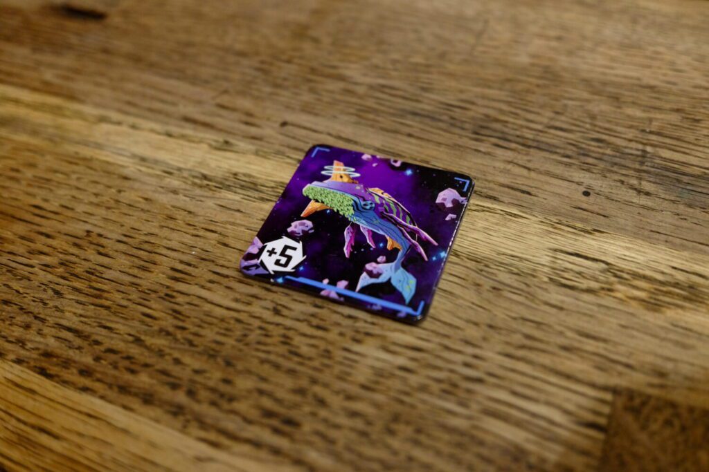 A single cardboard tile on a wooden table. The tile shows an illustration of a whale-like creature swimming/flying through deep space.