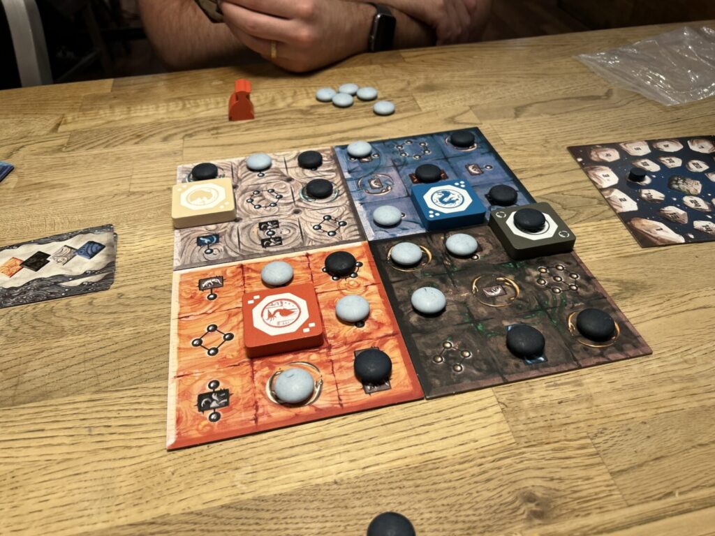Azure mid-game. The board is made up of four squares, in white, blue, orange, and black. Each is a 3 x 3 grid. Many of the spaces are covered with light blue or black stones. One space on each board is covered with a large wooden token, a mountain.