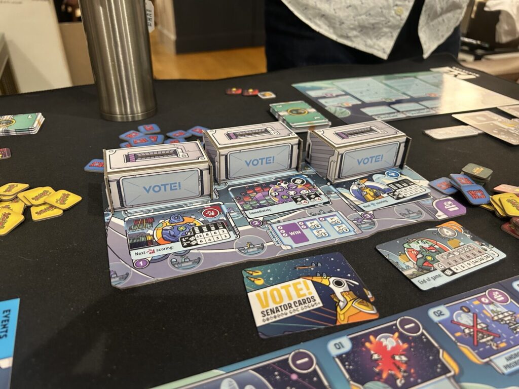Three cardboard cubes labelled "VOTE" sit in a row on a board. Three Senator cards sit in front of them.