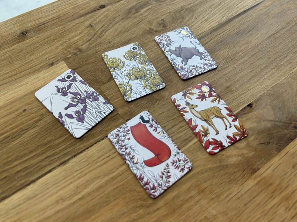 Five hanafuda tiles sit on a wooden table. Several tiles include embossed golden icons.
