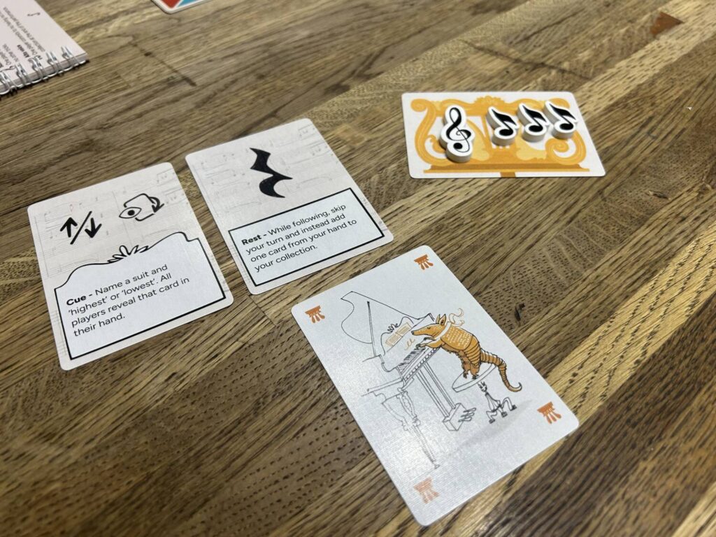 A player's power card, rest card, musician card, and music stand card, the latter with note tokens on it, sit on a wooden table.