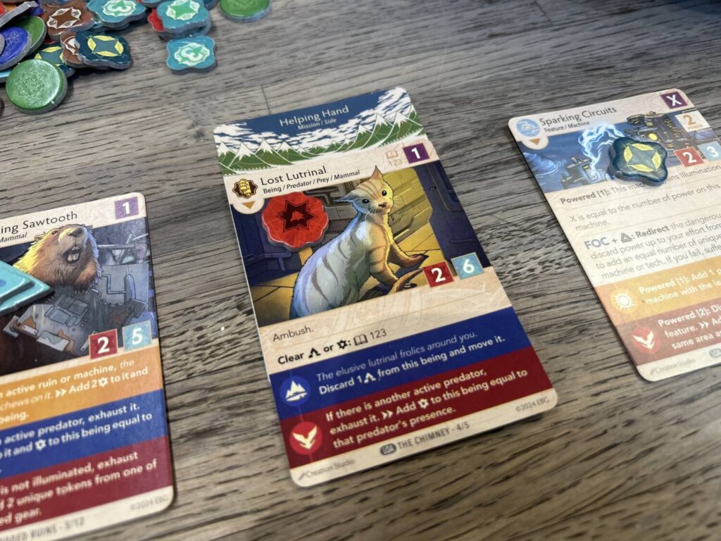 A close-up photograph of one card with some tokens on it, a mission card behind it, and other cards (with tokens) on each side.