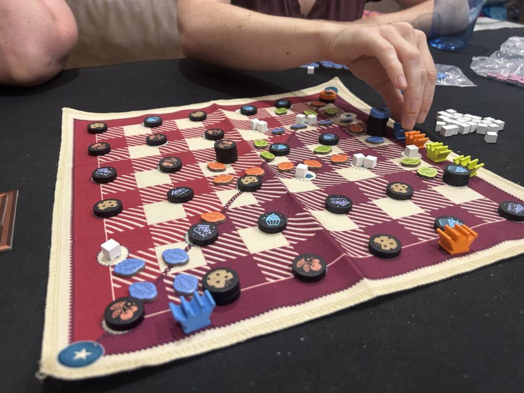 A gingham-style playmat with wooden crowns, wooden food tokens, and cardboard ant tokens.